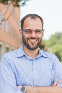 portrait of smiling man in light blue button-down shirt outdoors, backed by brick building